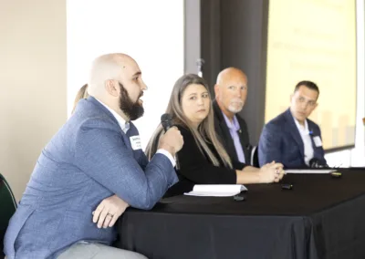 A man speaks into a microphone while sitting at a table during a panel discussion. Three other people, two men and a woman, sit attentively beside him. They are in a conference room with a presentation screen in the background.