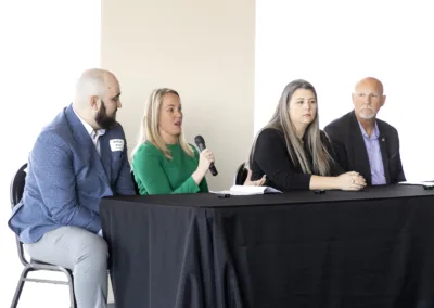 Four people sit at a table during a panel discussion. One woman, wearing a green top, speaks into a microphone. The group appears engaged in conversation, with two men and another woman at the table.