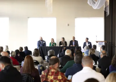 A panel of five people sit at a long table in front of an audience during a conference or seminar. The room is well-lit with large windows, and attendees are seated at round tables, listening to the speakers.