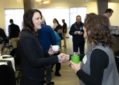 Two women are shaking hands at an indoor event. One holds a coffee cup, and the other a green tumbler. People are mingling in the background, and tables are set up for the event.