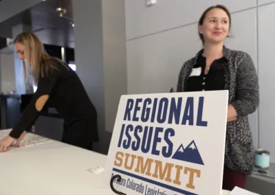 Two individuals are at a registration table. One is setting up name tags, while the other stands next to a sign that reads "Regional Issues Summit" with mountain graphics. The background shows a modern interior setting.