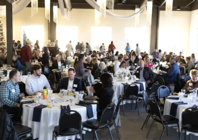 A large group of people seated around tables in a brightly lit banquet hall, engaged in conversation. Tables are set with white tablecloths, blue napkins, and glassware. A decorated Christmas tree is visible in the background.