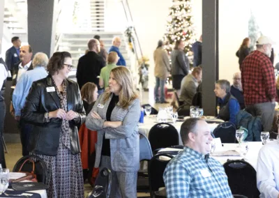 A group of people gathered in a spacious room for a holiday event. Two women are conversing in the foreground, while others sit at tables. A decorated Christmas tree and staircase are visible in the background.