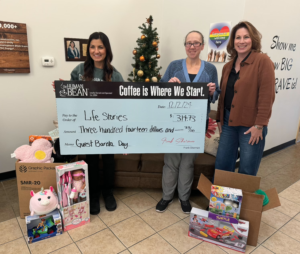 Three people stand indoors holding a large check for $314.73 made out to "Life Stories." They are surrounded by boxes of toys. The check notes "Guest Barista Day" and is dated 12/12/24. A "Coffee is Where We Start" sign is visible.