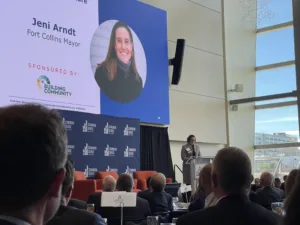 A speaker is addressing an audience at a conference. A large screen displays a portrait of Jeni Arndt with the text "Fort Collins Mayor" and sponsor logos. Attendees are seated and focused on the presentation.