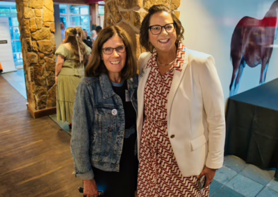 Two women smile for a photo indoors. One wears glasses, a denim jacket, and a black dress, while the other wears glasses, a patterned dress, and a white blazer. People and a stone wall are visible in the background.