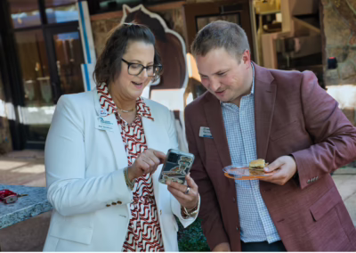 Two people in business attire stand outside. The woman shows something on her phone to the man, who holds a plate with food. They both appear engaged and smiling, with a horse silhouette visible in the background.