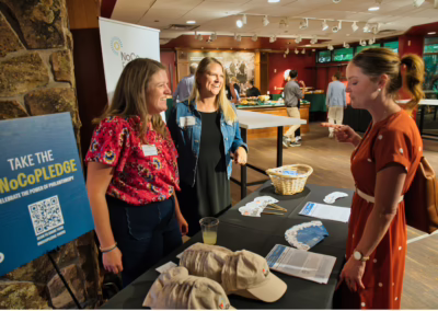 Three women chat at an indoor event table covered with hats, flyers, and a basket. A sign reads “Take the #NoCoPledge.” Other attendees are seen mingling in the background.