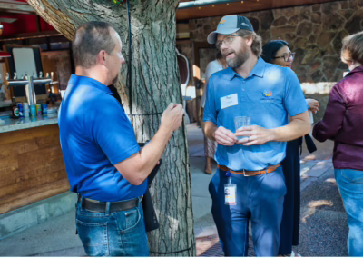 Two men stand by a tree, having a conversation at an outdoor event. One man holds a drink; both wear blue shirts and name tags. Other people and a bar area are visible in the background.