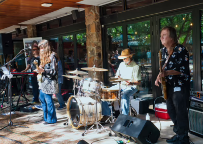 A four-piece band performs outdoors, featuring a keyboardist, guitarist, drummer, and bassist, with sunlight streaming in and people visible through large glass windows behind them.