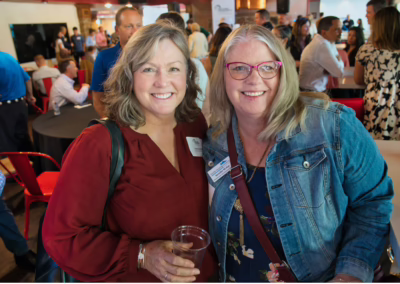 Two smiling women pose together at a busy indoor social event. One holds a drink and wears a red top, while the other wears glasses, a denim jacket, and a conference badge. People mingle in the background.