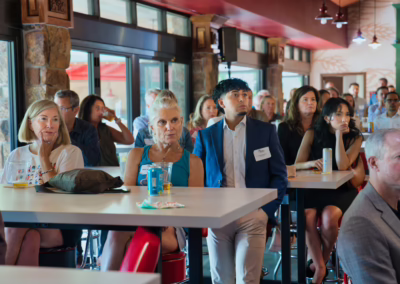A diverse group of people sit and stand at tables in a bright room, attentively listening to a speaker out of frame. Some hold drinks or snacks, and large windows let in natural light.