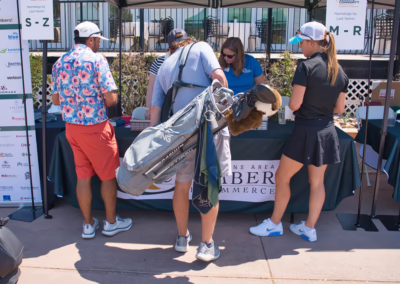 Four people stand at an outdoor registration table under a tent at a golf event. Two women assist two men, one carrying a golf bag. Event sponsor banners are displayed on the left.