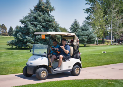 Two men ride in a white golf cart on a paved path at a golf course, surrounded by green grass and trees under a clear blue sky.
