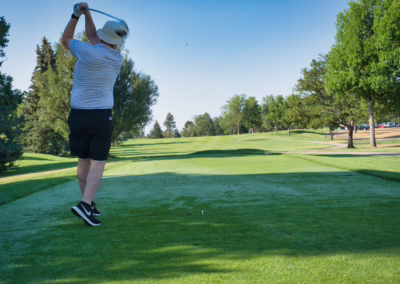 A person wearing a hat, patterned shirt, and shorts swings a golf club on a lush green golf course, with trees and a clear blue sky in the background.