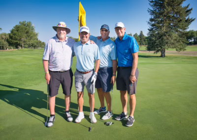 Four men stand together on a golf course green, smiling at the camera. They are dressed in golf attire with clubs, and a yellow flag is visible behind them. The weather is sunny with trees in the background.