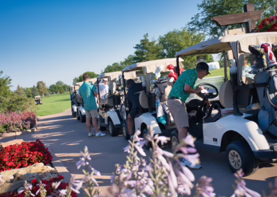 Several people in golf attire stand by a row of golf carts on a paved path beside a colorful flower bed, preparing for a day on the green under a clear blue sky.