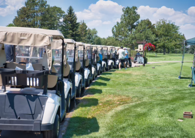 A row of golf carts parked on a golf course, with people gathered near a tent in the background under a sunny sky and green trees.