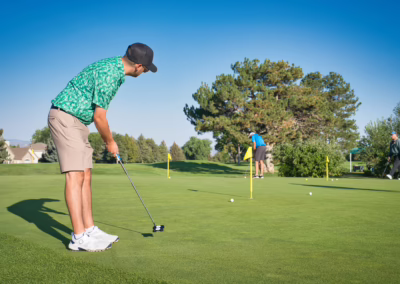 A man in a green patterned shirt and shorts prepares to putt on a golf green while two other golfers play nearby under a clear blue sky, surrounded by trees and manicured grass.