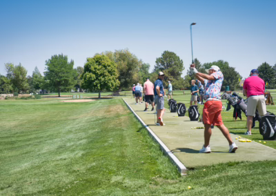 People practice golf swings at a driving range on a sunny day, with golf bags and balls on the ground. Trees, grass, and blue sky are visible in the background.