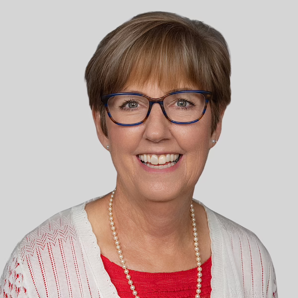 Smiling woman with short light brown hair, wearing glasses, a red top, a white sweater, and a pearl necklace, posing against a plain light gray background.