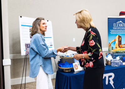 Two women shake hands in front of a table with promotional materials and posters at an indoor event or conference. One woman wears a blue shirt and white pants; the other wears a black floral top.