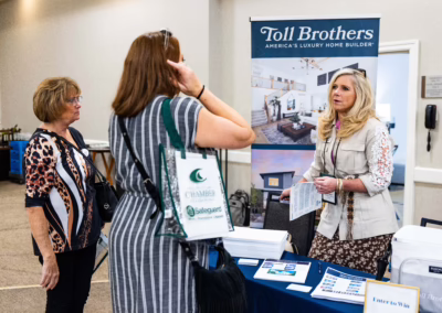 A woman stands behind a table with brochures, speaking to two women at a Toll Brothers home builder booth during an indoor event or expo.