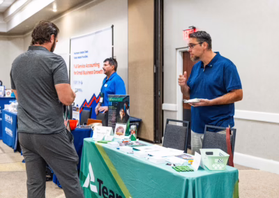 Two men talk at a business expo, standing near a booth with informational materials on a green tablecloth. Other vendors and displays line the background in a well-lit conference room.