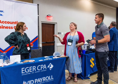 Three people stand and talk at a business expo; two women are behind a table with an “Eger CPA” and “Avid Payroll” banner, while a man stands in front, holding brochures and engaging in conversation.