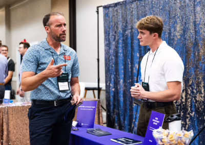 A man in a blue shirt gestures while talking to a young man in a white shirt at a booth with brochures and giveaways during an indoor event or conference. Other people and booths are visible in the background.