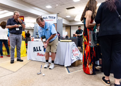 A man in casual clothes putts a golf ball indoors at a business expo while others stand nearby, talking and observing various booths with banners and products.