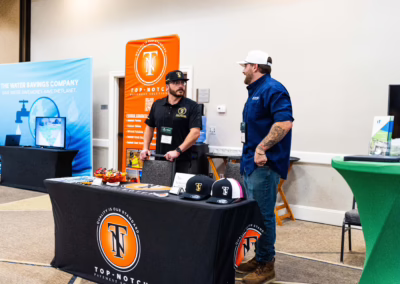 Two men stand behind a promotional table with branded hats and marketing materials at a business expo. Banners for "Top-Notch" and "The Water Savings Company" are displayed in the background.