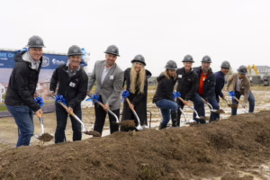 A group of people in hard hats and coats use shovels to break ground at a muddy construction site, smiling for the camera during a rainy day. A sign and partially built homes are visible in the background.