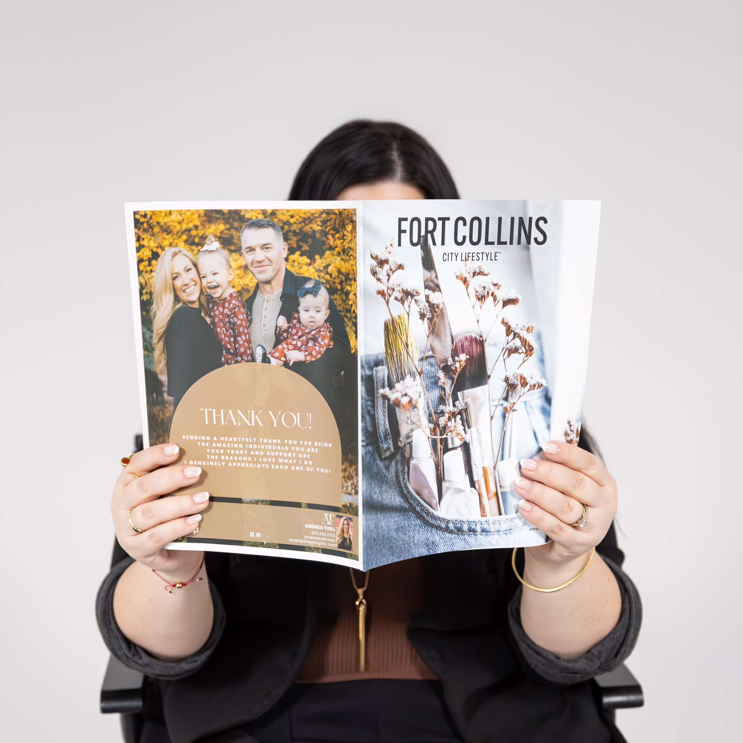 A person with dark hair holds up and reads a “Fort Collins City Lifestyle” magazine, obscuring their face. The magazine's cover shows dried flowers in vases, and an inside page features a family portrait and a thank you note.