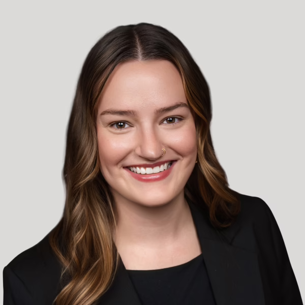 A woman with long brown hair, wearing a black blazer and top, smiling at the camera. She has a nose ring and is posed against a plain light gray background.