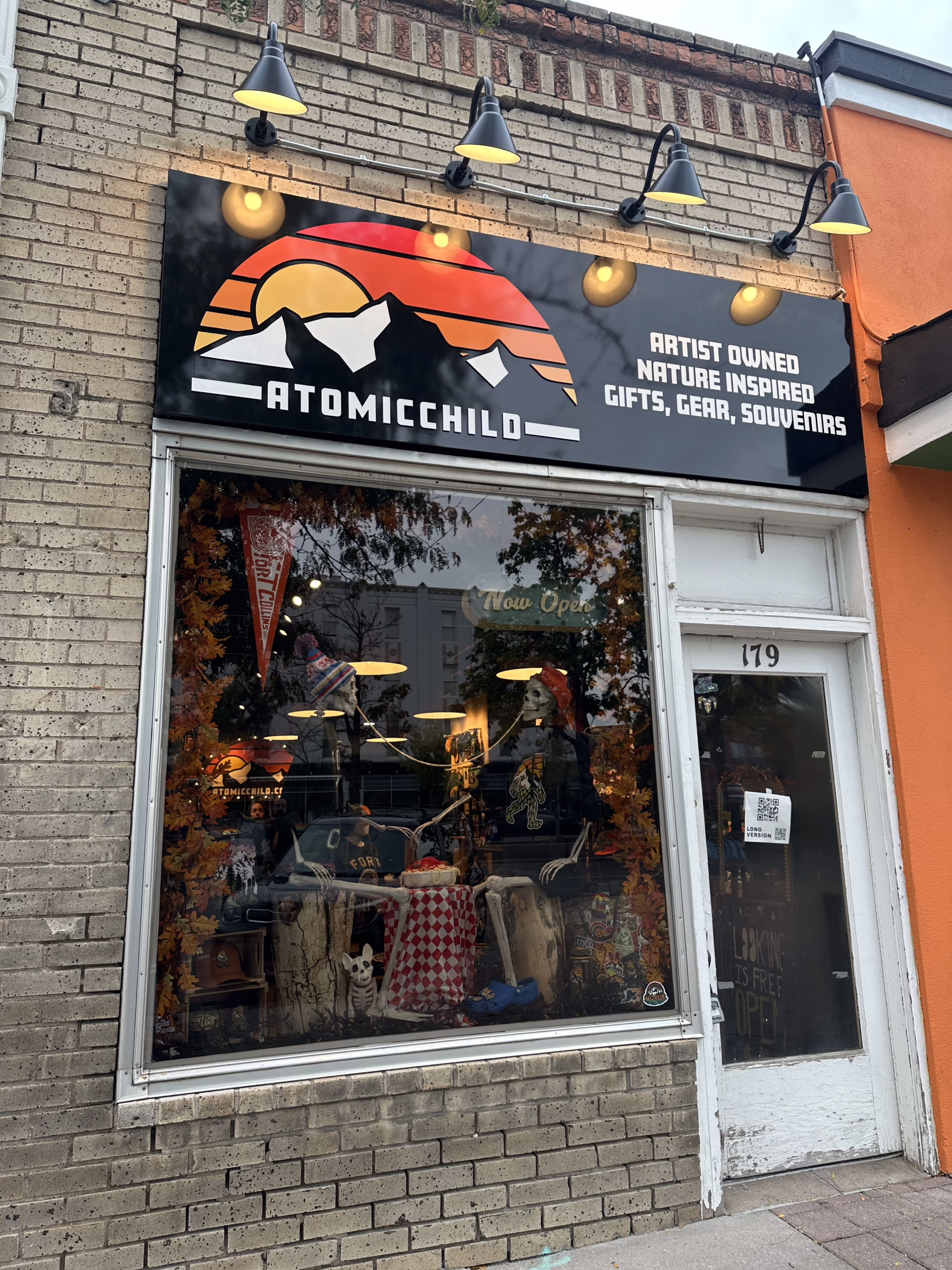 Storefront of "Atomicchild," featuring a sign with a stylized mountain and sun logo. The window displays nature-inspired gifts and gear, with a "New Open" sign. The brick building has five lights above the sign.