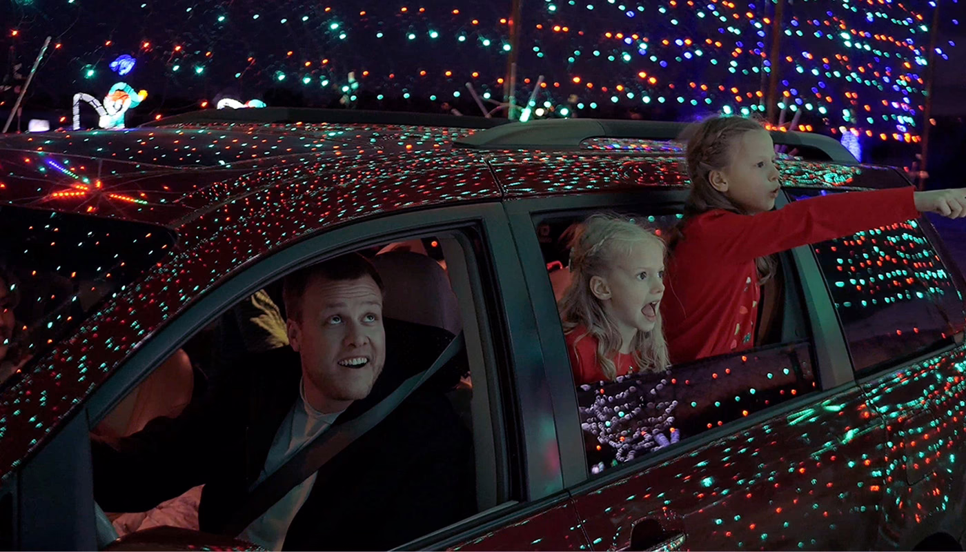 A man drives a car while two children lean out of the back window, excitedly pointing and looking at colorful holiday lights covering the car and the surroundings.