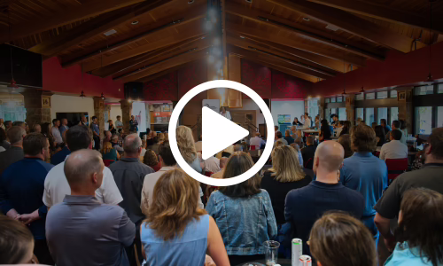 A large group of people gather indoors, listening to a speaker at the front of the room. The setting is well-lit with wooden ceilings. A play button icon is overlaid in the center, suggesting a video.