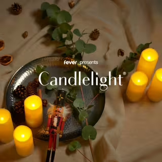 A metal tray with a nutcracker, pinecones, and greenery sits on beige fabric, surrounded by five glowing candles. The text "fever presents Candlelight" is centered over the image.
