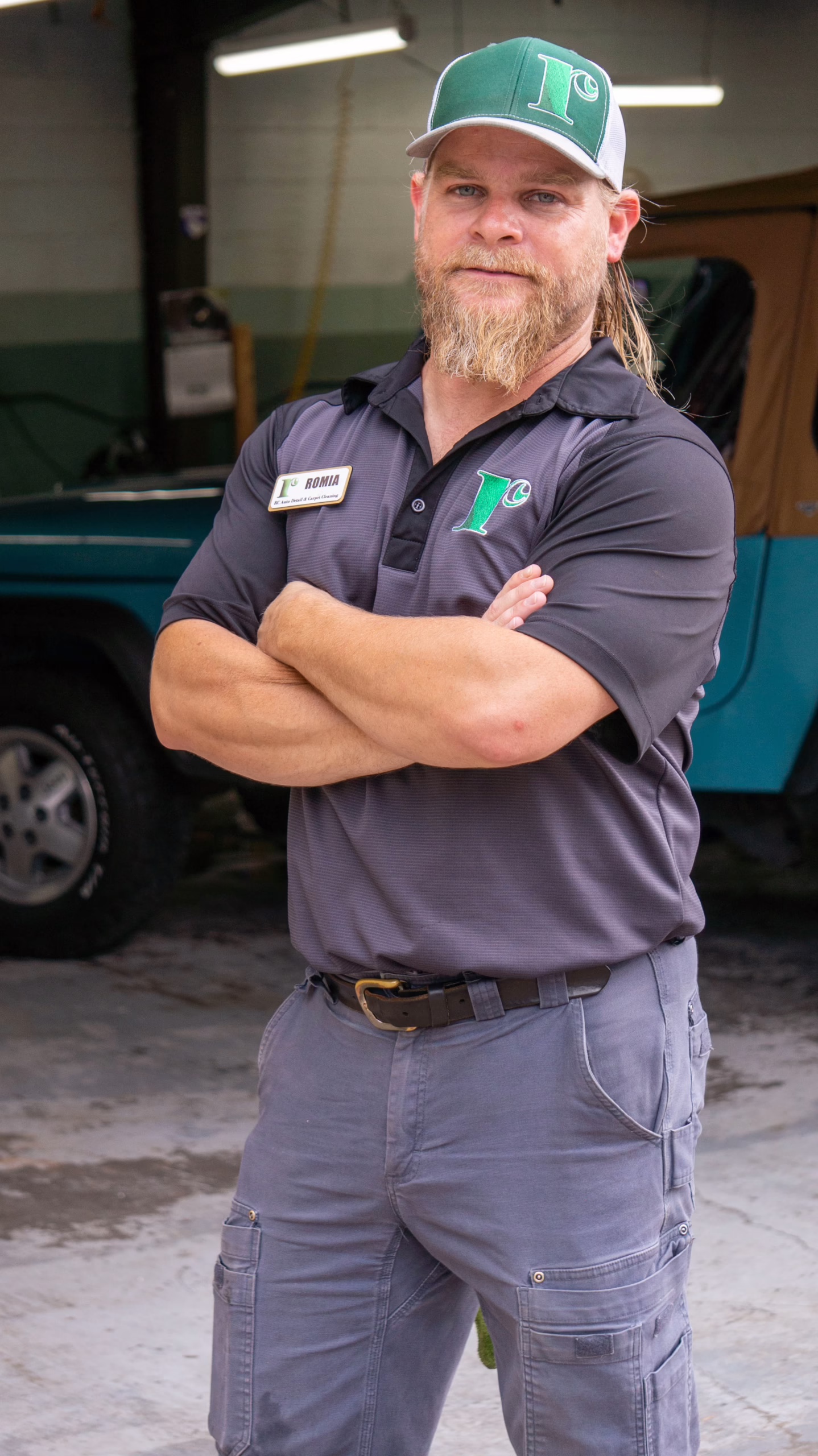 A man with a beard and long hair, wearing a green and white cap, dark polo shirt, and cargo pants, stands with arms crossed in a garage; a blue vehicle is visible in the background.