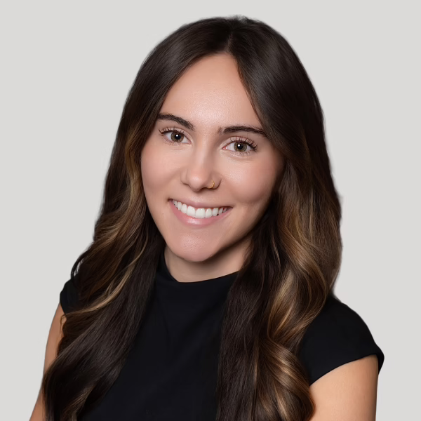 A woman with long, wavy brown hair and a nose ring smiles at the camera. She is wearing a black top and is posed in front of a plain, light gray background.