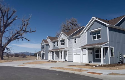 A row of modern, gray and white two-story townhouses with attached garages stands along a quiet street. A large leafless tree and open field are visible in the background under a clear blue sky.