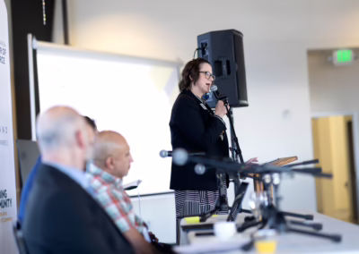 A woman speaks at a podium with a microphone during a panel discussion, while three men sit and listen in the foreground. A large screen and microphones are visible in the brightly lit room.