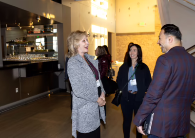 Three people stand and converse in a modern, well-lit indoor space near a bar area. Two women and one man, all wearing conference badges, are smiling and engaged in conversation.