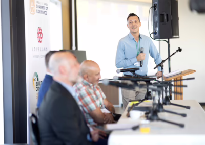 A man stands at a podium speaking into a microphone, smiling at the audience. Three panelists sit at a table in the foreground, with microphones in front of them. Banners from local organizations are visible in the background.