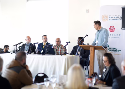 A man stands at a podium speaking to a seated panel of five men at a conference, with microphones and laptops on the table. Audience members sit at round tables in the foreground. Chamber of Commerce banners are visible in the background.