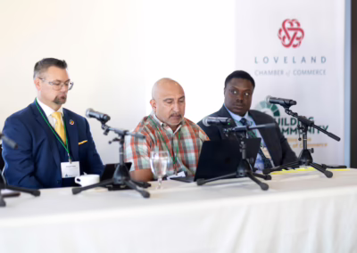 Three men sit at a table with microphones and a laptop during a panel discussion. Two banners behind them display the Loveland Chamber of Commerce and Wilds & Commons logos.