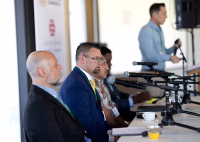 Four men sit side by side at a panel table with microphones, while a fifth man stands in the background near a podium. They appear to be participating in a conference or discussion event.