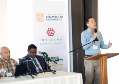 A man speaks at a podium with a microphone during a conference, while two other men sit at a table nearby. Banners for local chambers of commerce are visible in the background.