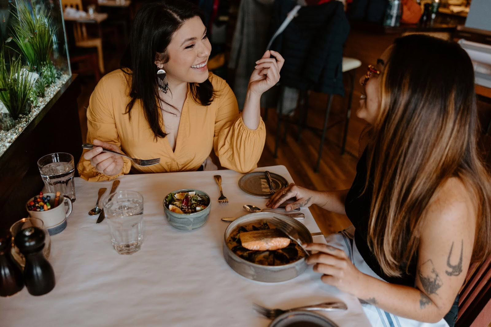 Two women sit at a restaurant table, smiling and talking while enjoying their meals. Both have plates of food in front of them, and the atmosphere appears casual and friendly.